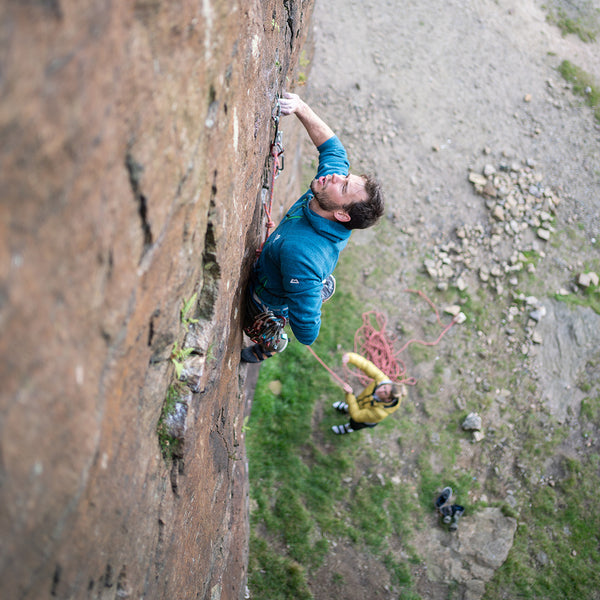 A climber on a rope high on a rock face contemplates his next move as his belay partner watches from the ground.