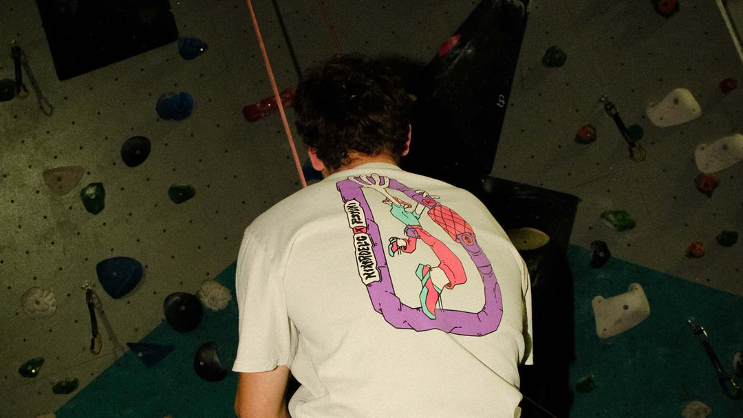 A climber in an off white climbing t-shirt featuring a purple carabiner and mushroom headed climber design, hangs from a rope at Parthian Climbing Centre in Manchester